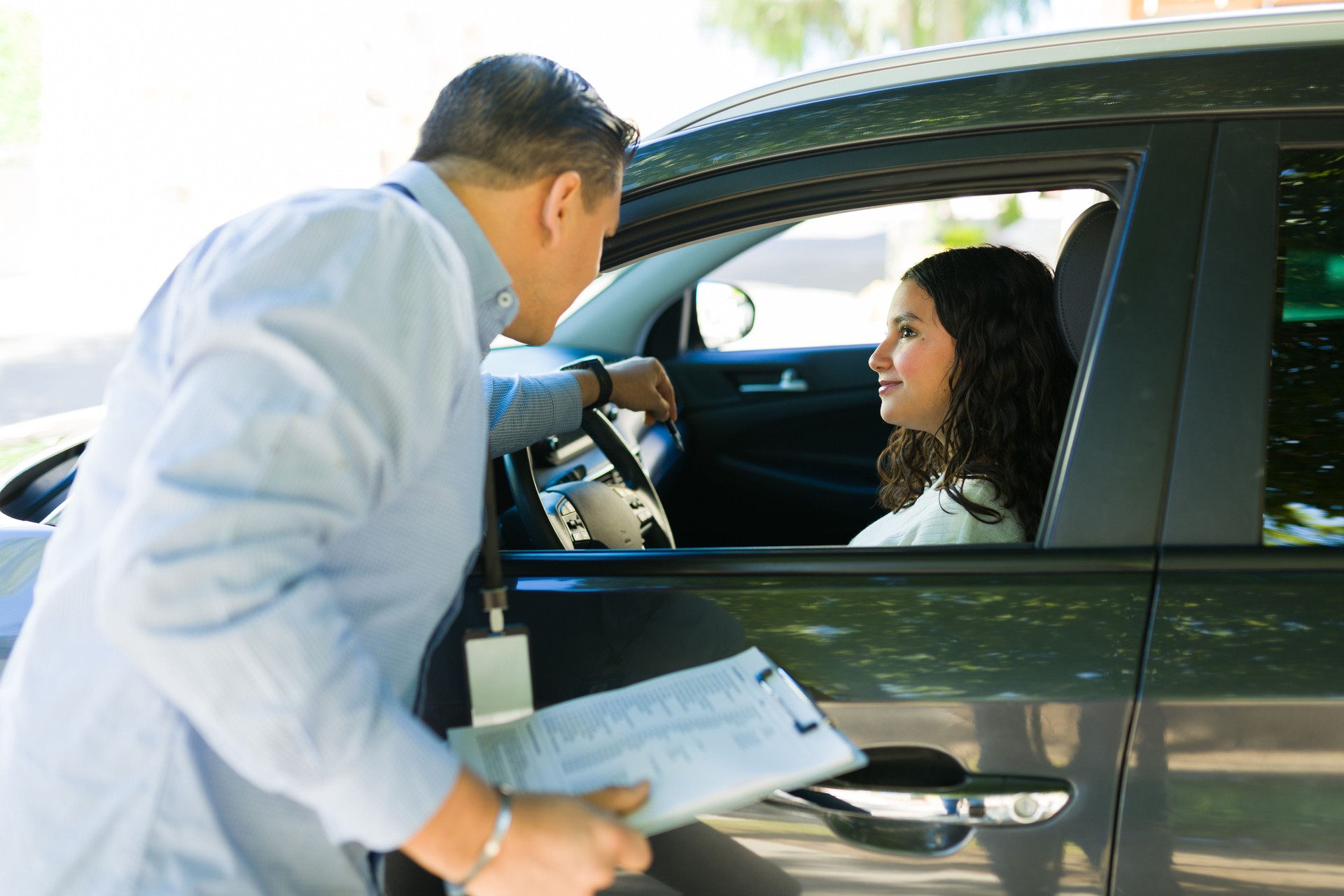 Hispanic driving instructor providing guidance to a teenage girl during a driving lesson while holding a clipboard